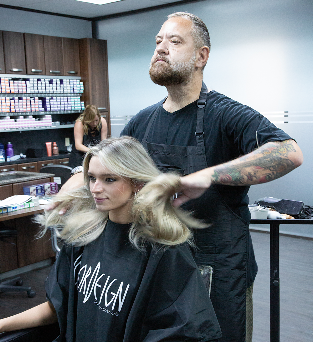 A stylist with tattooed arms works on a woman's hair in a salon, with shelves of hair products visible in the background.