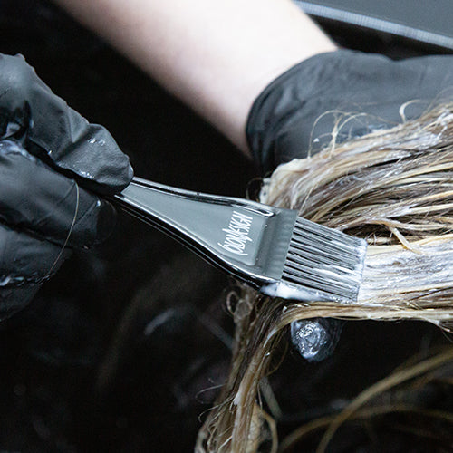 Close-up of a stylist's applying hair color with a brush labeled 'ColorDesign' to a client's hair.