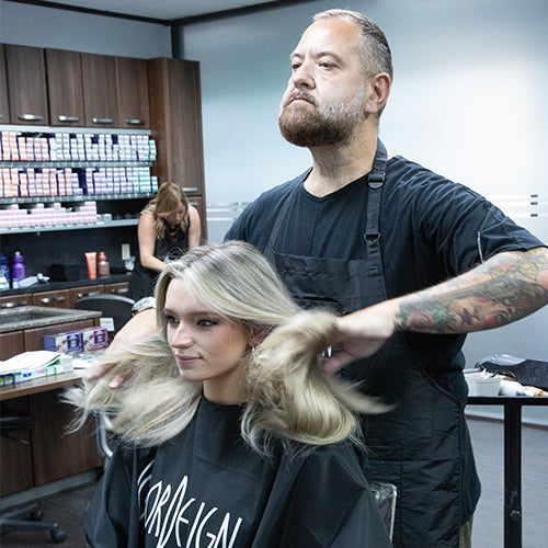 A stylist with tattooed arms works on a woman’s hair in a salon, with shelves of hair color products visible in the background.