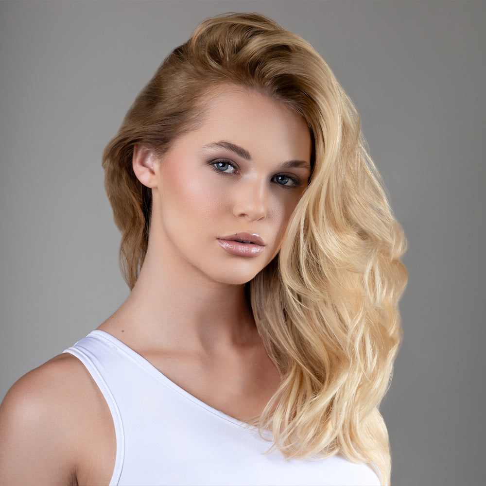 Portrait of a woman with voluminous blonde hair styled in loose waves, wearing a white top against a light gray background.